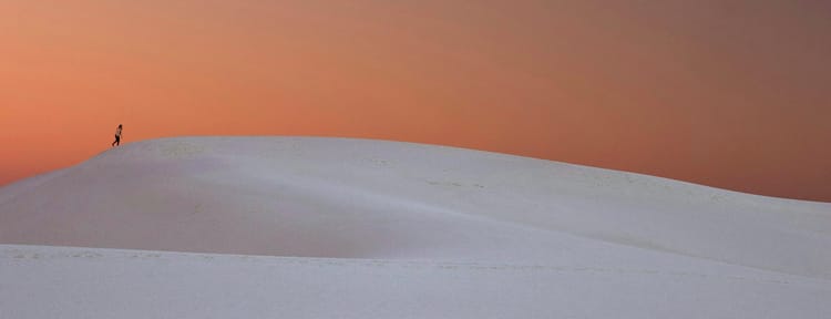 person in the far distance walking over a hill of sand under an empty pale orange sky