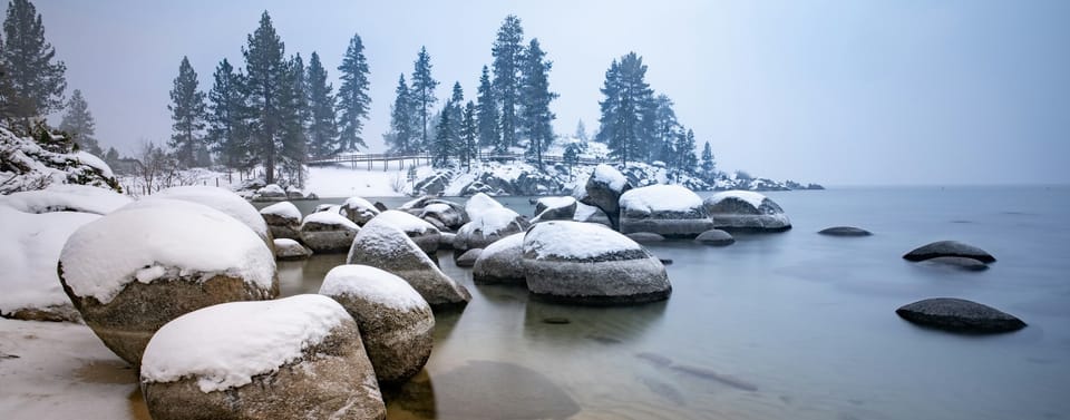 snow-covered boulders at water’s edge with trees in the distance